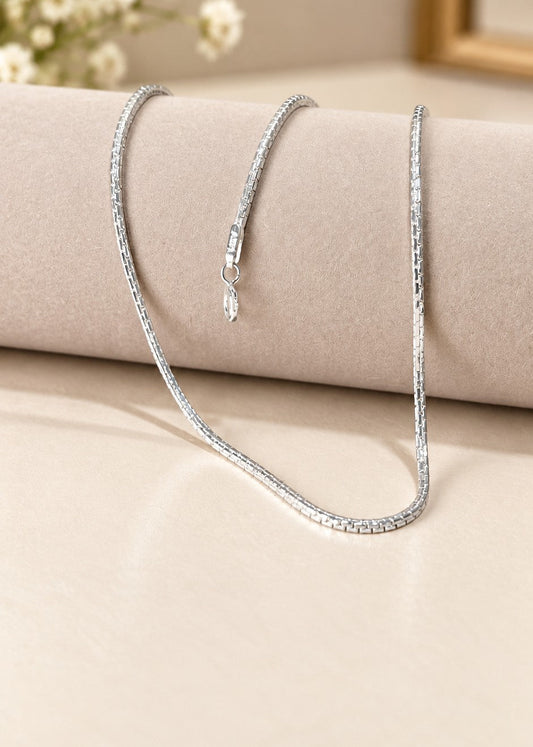 A silver-toned tennis chain necklace draped over a beige cylindrical jewelry display, with a small pendant clasp visible and a softly blurred neutral background with small white flowers.